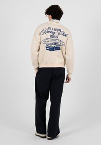 Personne aux cheveux bouclés portant un pull beige avec le texte "Racing Club Berlin" et un motif de voiture, associée à un pantalon noir et des chaussures blanches.