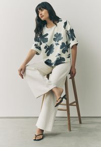 White floral-print blouse with black flowers, paired with wide-legged cream pants and black flip-flops, seated on a wooden stool.