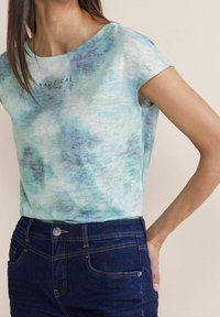 Kvinna som bär en ljusblå mönstrad t-shirt med kort ärm som har texten "NAUTICAL" samt mörkblå jeans, med handen vilande på höften.