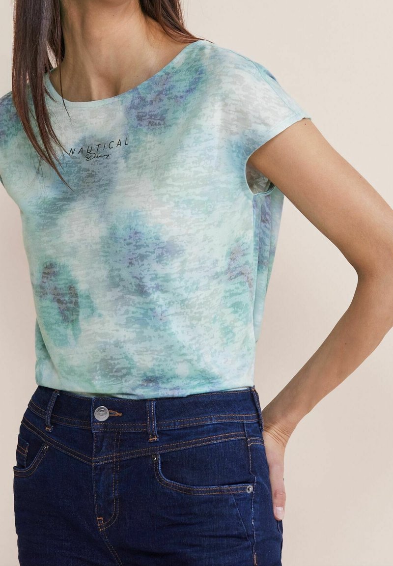 Kvinna som bär en ljusblå mönstrad t-shirt med kort ärm som har texten "NAUTICAL" samt mörkblå jeans, med handen vilande på höften.