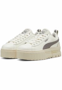 Vit sneaker tillverkad av slät syntetiskt material, med en tjock, räfflad sula, grå detaljer och en logotyp på tungan och sidan.