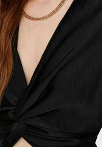 Top nero con un design annodato sul davanti, realizzato in un tessuto liscio. Una collana a catena in oro rosa è visibile al collo.