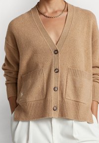 Brun stickad cardigan med V-krage, framknappar, två stora fickor och ribbade muddar och nederkant. Har en liten logotyp på den vänstra fickan.