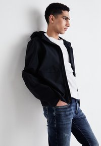 Black waterproof jacket with a hood, zip closure, and front pockets. Paired with a white shirt and dark blue distressed jeans.