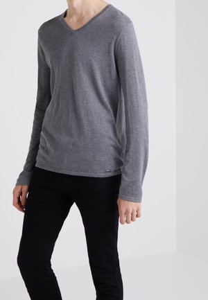 Personne portant un pull gris à manches longues en V et un pantalon noir, debout devant un fond clair uni.
