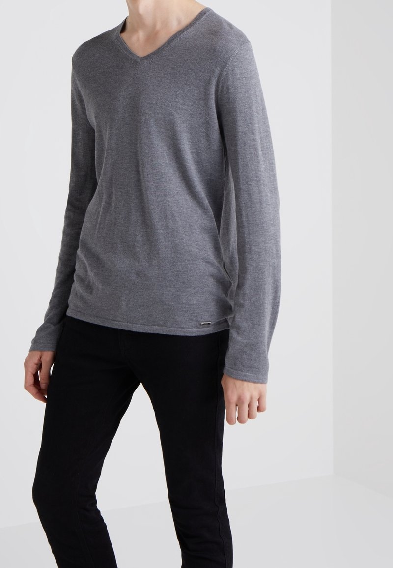 Personne portant un pull gris à manches longues en V et un pantalon noir, debout devant un fond clair uni.