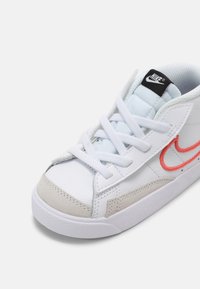 Baskets basses blanches Nike avec lacets blancs, embout gris clair, swoosh rouge brodé et étiquette noire sur la languette.