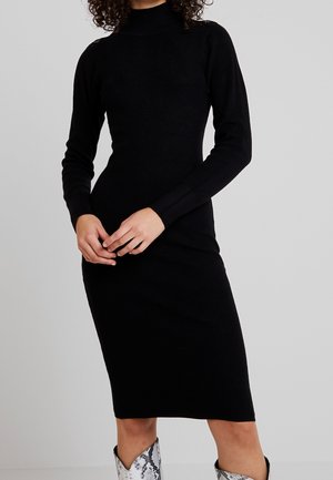 Femme portant une robe midi noire ajustée à manches longues et des bottes argentées à motif peau de serpent, posant les mains croisées devant elle.