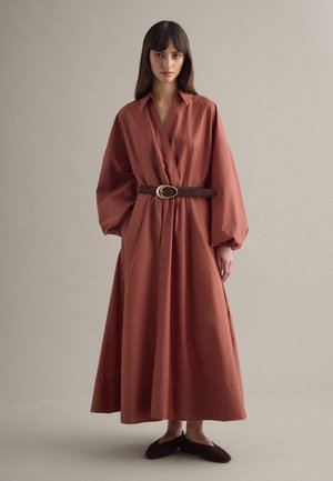 Mujer con un vestido color oxidado, de manga larga y largo hasta el tobillo, con un cuello ancho y un cinturón marrón oscuro con hebilla dorada.