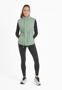 Light green sleeveless vest with a high collar, drawstring waist, and diamond quilted pattern, paired with black leggings and running shoes.