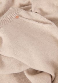 Pull en tricot beige fabriqué à partir d'un matériau doux. Il présente un col en côtelé à motif et un petit logo rond près de l'épaule.