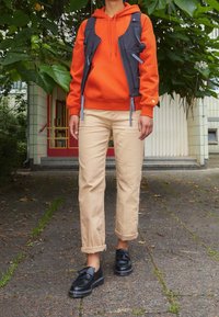 Persona de pie al aire libre vistiendo una sudadera naranja, un chaleco utilitario negro, pantalones beige arremangados y mocasines negros con borlas en un camino pavimentado.