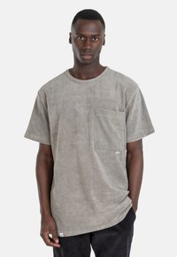 Grau meliertes T-Shirt mit einer Brusttasche, Rundhalsausschnitt, kurzen Ärmeln und einem lockeren Schnitt. Das Material wirkt weich und leicht flauschig.