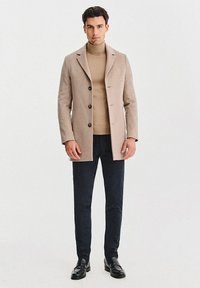 Beżowy wełniany płaszcz z wyciętym kołnierzem, trzema guzikami, w zestawie z beżowym golfem i ciemnogranatowymi dopasowanymi spodniami, uzupełniony czarnymi mokasynami.