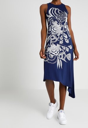 Marinblå ärmlös klänning med vitt blommönster, med asymmetrisk fåll och mjukt, slätt tyg; bärs med vita sneakers.