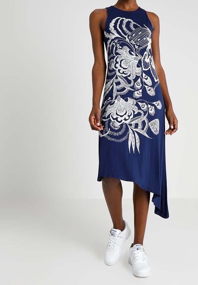 Granatowa sukienka bez rękawów z białym kwiecistym wzorem, z asymetrycznym dołem i miękkiego, gładkiego materiału; noszona z białymi trampkami.