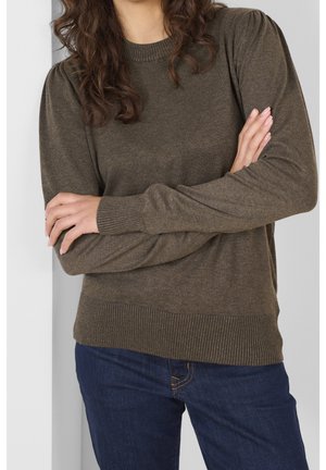 Femme les bras croisés portant un pull en maille marron à manches longues et un jean bleu foncé, debout sur un fond clair.