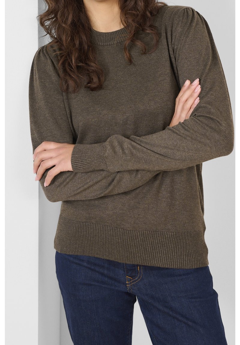 Femme les bras croisés portant un pull en maille marron à manches longues et un jean bleu foncé, debout sur un fond clair.