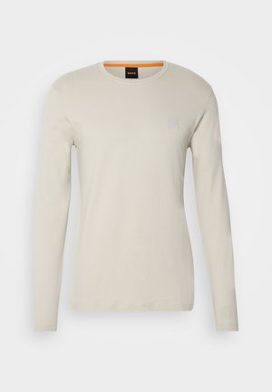 Chemise en coton beige clair à manches longues, avec un col rond et un petit logo carré accentué en gris sur la poitrine.