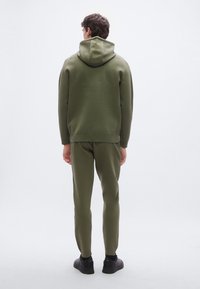 Sudadera con capucha verde oliva y pantalones a juego, hechos de tela suave con detalles acanalados. Capucha en la parte trasera, diseño ajustado, combinados con zapatos negros.