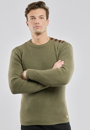 Ung mand med armene over kors iført en olivengrøn ribstrikket sweater med fire brune knapper på venstre skulder mod en ensfarvet baggrund.