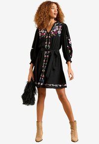 Robe noire avec broderies florales dans des couleurs comme le rose et le bleu, présentant un décolleté en V, de longues manches bouffantes et une taille nouée.