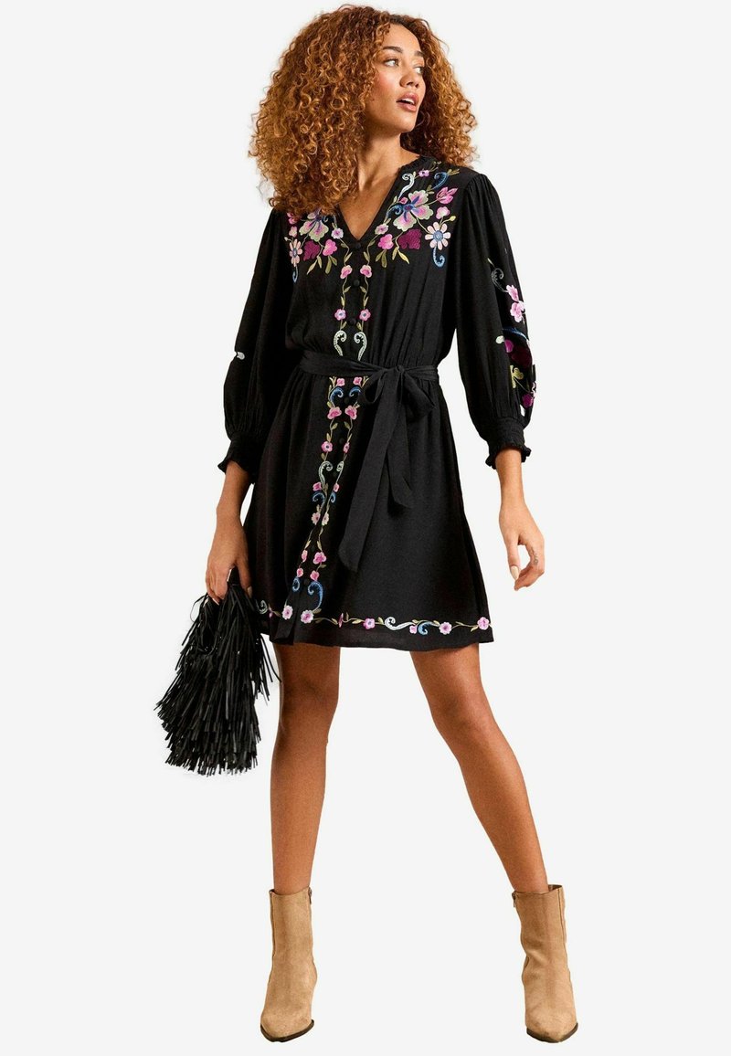 Robe noire avec broderies florales dans des couleurs comme le rose et le bleu, présentant un décolleté en V, de longues manches bouffantes et une taille nouée.