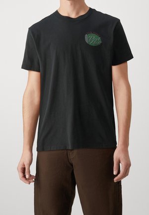 Mann trägt schwarzes kurzärmeliges T-Shirt mit kleinem grün-rotem "New York Pool Hall" Logo und braune Hose vor einfarbigem Hintergrund.