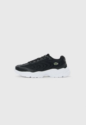 Baskets de sport noires avec une tige texturée, semelle épaisse blanche, lacets noirs et petit logo de crocodile vert sur le côté.