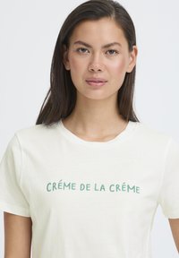 Kremno bela bombažna majica z napisom "CRÉME DE LA CRÉME" v zeleni vezenini. Klasičen kroj z okroglim izrezom. Gladka tekstura.