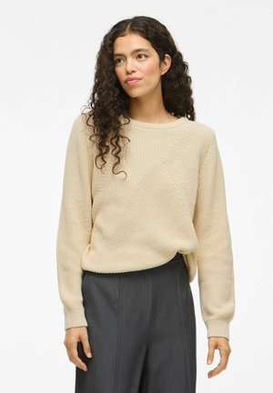 Beige knitted sweater with long sleeves, round neckline, and textured patterns. Paired with gray high-waisted trousers.