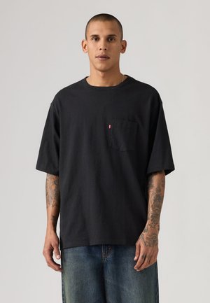 Moški s kratkimi lasmi in tetovažami, oblečen v ohlapno črno majico z žepi in modre kavbojke, stoji pred svetlim enobarvnim ozadjem.