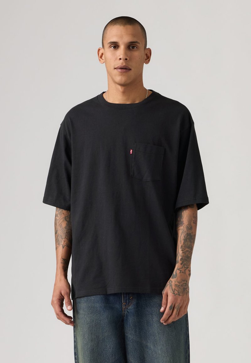 Άντρας με κοντά μαλλιά και τατουάζ, που φοράει χαλαρό μαύρο t-shirt με τσέπες και μπλε τζιν, στέκεται μπροστά σε απλό φωτεινό φόντο.