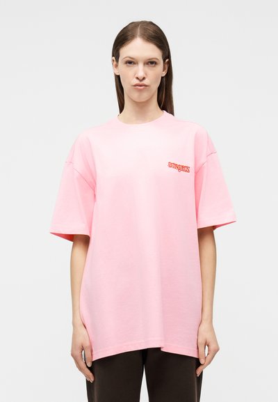 Γυναίκα με μακριά καστανά μαλλιά που φοράει ένα φαρδύ ροζ ανοιχτό t-shirt με κόκκινο κείμενο στο αριστερό στήθος, στέκεται μπροστά σε απλό λευκό φόντο.