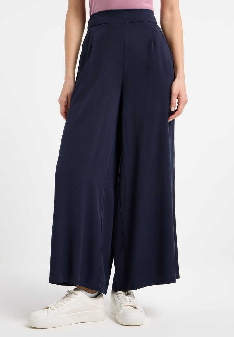 Pantaloni larghi navy con vita alta e pieghe frontali, realizzati in un tessuto liscio. Combinati con sneakers bianche.