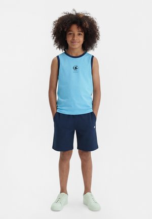 Enfant aux cheveux bouclés portant un débardeur bleu clair, un short bleu marine et des baskets blanches, debout les mains dans les poches.