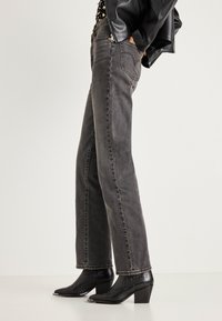 Άτομο που φοράει στενά μαύρα τζιν παντελόνια σε ίσια γραμμή με ξεθωριασμένη όψη, μαύρα μποτάκια με τακούνι και μυτερή μύτη, και μαύρο δερμάτινο μπουφάν, έχοντας τα χέρια στις τσέπες.