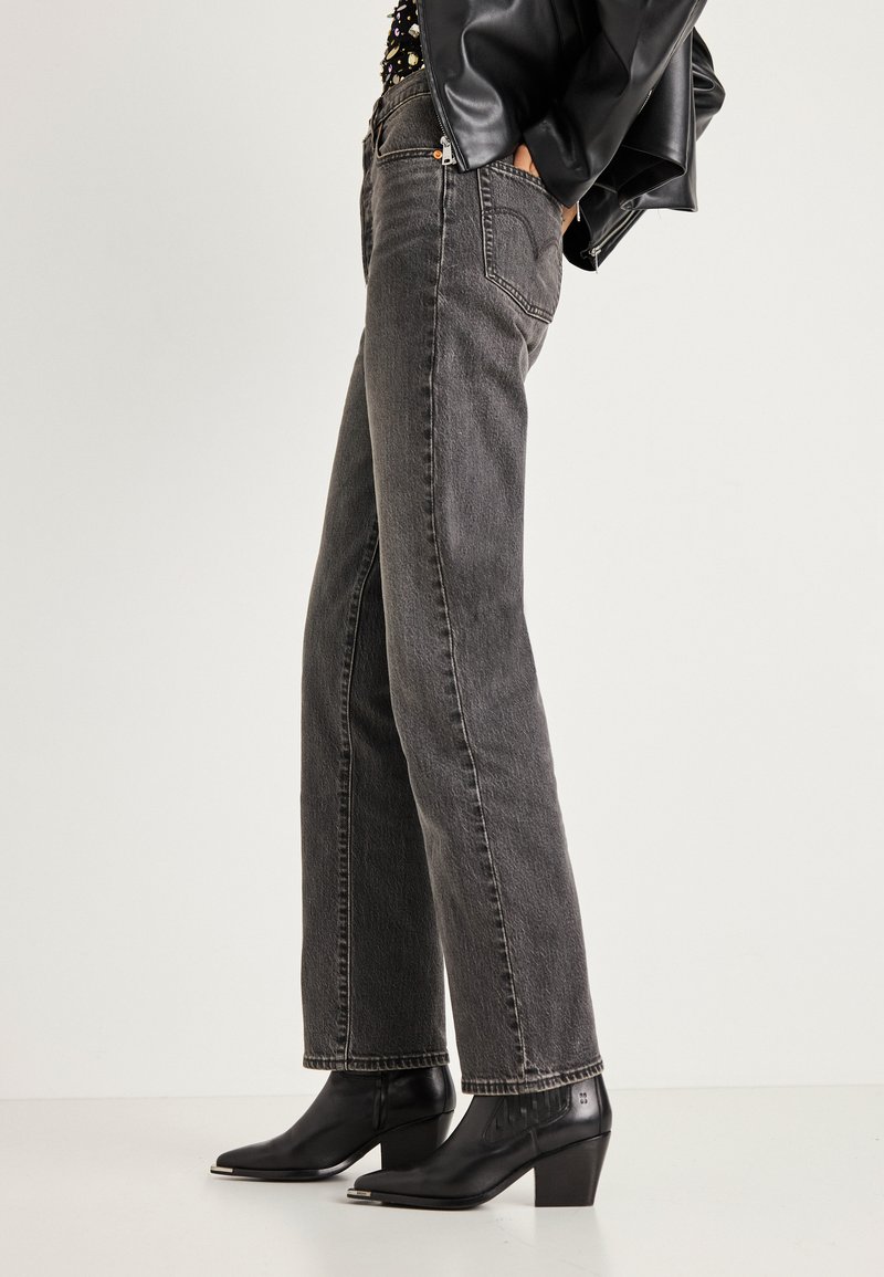 Άτομο που φοράει στενά μαύρα τζιν παντελόνια σε ίσια γραμμή με ξεθωριασμένη όψη, μαύρα μποτάκια με τακούνι και μυτερή μύτη, και μαύρο δερμάτινο μπουφάν, έχοντας τα χέρια στις τσέπες.