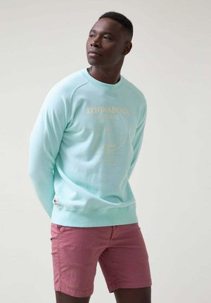Sudadera azul claro con estampado dorado, puños y bajo acanalados. Combinada con pantalones cortos rosas, de textura suave y corte relajado. Detalle de marca sencillo.