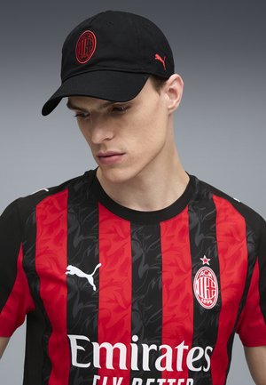 Młody mężczyzna w czarnej czapce Puma i czerwono-czarnej koszulce piłkarskiej AC Milan z logo sponsora Emirates, spoglądający w dół.