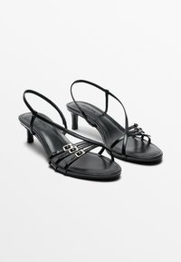 Paire de sandales à talons noires avec fines brides et petites boucles argentées sur fond blanc.