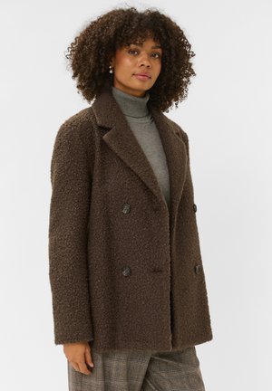 Femme aux cheveux bouclés portant un manteau marron texturé à double boutonnage, un col roulé gris et un pantalon à carreaux sur un fond blanc uni.