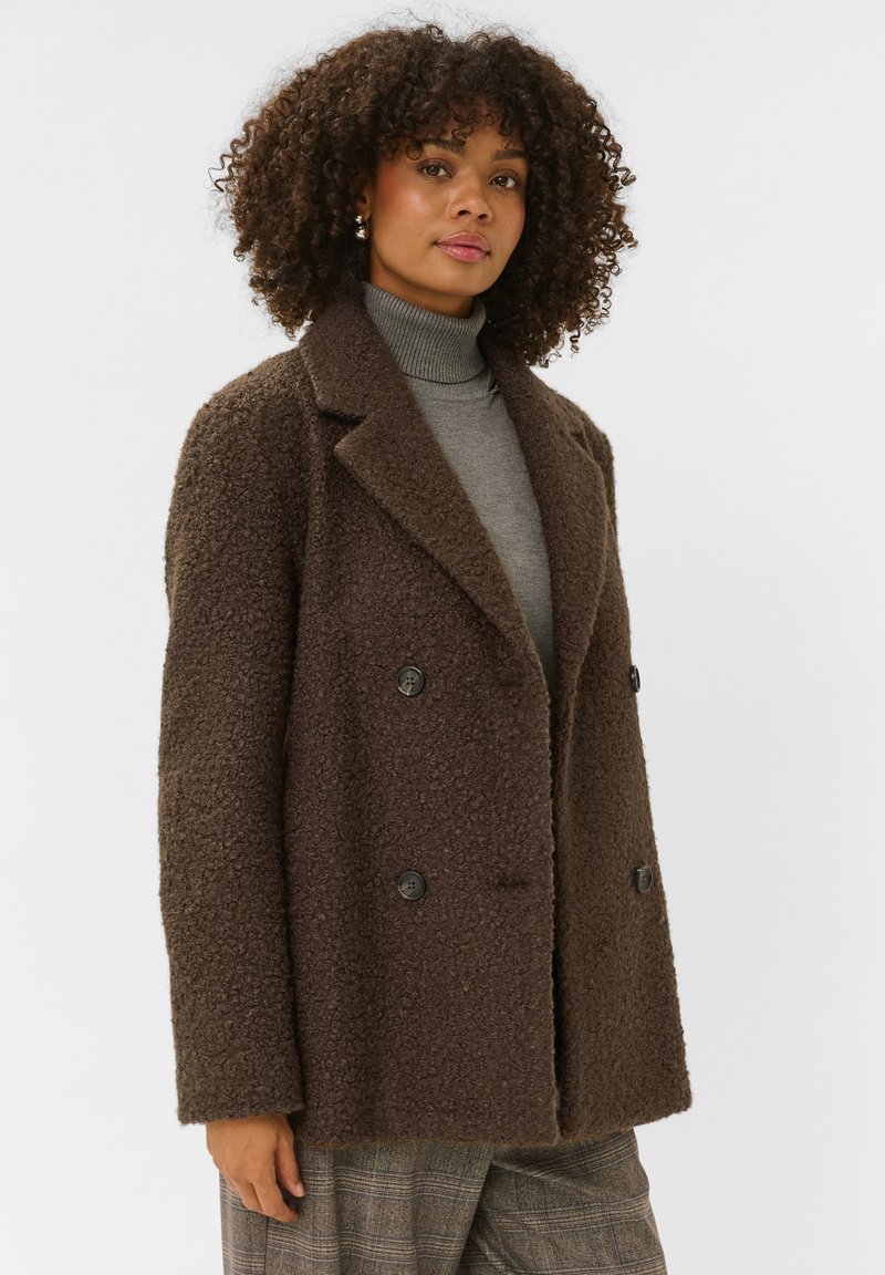 Femme aux cheveux bouclés portant un manteau marron texturé à double boutonnage, un col roulé gris et un pantalon à carreaux sur un fond blanc uni.
