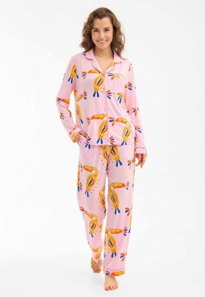 Kobieta uśmiechnięta, ubrana w różowy zestaw piżam z guzikami z kolorowymi nadrukami tukana, stoi boso na białym tle.