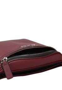 Bordeauxrood leren clutchtas met een getextureerde afwerking, voorzien van een zilveren rits en een dun leren label, gedeeltelijk geopend om een zwarte binnenkant te tonen.