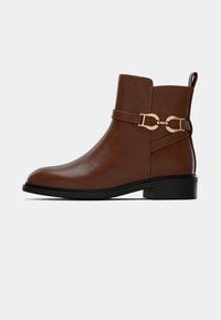 Bruine leren enkelboot met een gesloten neus en een lage blokhak. Heeft een decoratieve gouden gesp en riemdetail aan de zijkant.