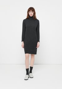 Ženska stoji oblečena v temno sivo žametno obleko s turtleneck izrezom, črnimi nogavicami in belimi natikači proti enobarvni beli ozadju.