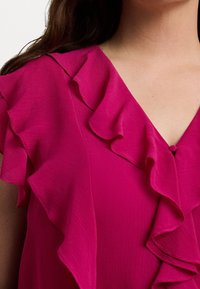 Fuchsia blus med volangkrage och axeldetaljer, med en texturerad tyg och en liten knäppning framtill.