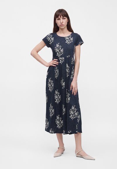 Femme portant une robe midi bleu marine avec des motifs floraux blancs, debout, une main sur la hanche et des chaussures plates beiges sur fond blanc.