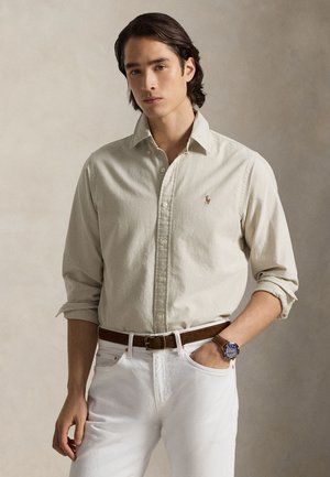 Chemise beige à boutons en coton, avec manches retroussées et logo sur la poitrine, associée à un jean en denim blanc et une ceinture marron.